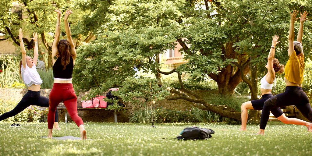 Vier Yogaschülerinnen praktizieren die Krieger 1 Pose (Virabhadrasana 1) im grünen Hofgarten Augsburg, umgeben von idyllischer Natur.