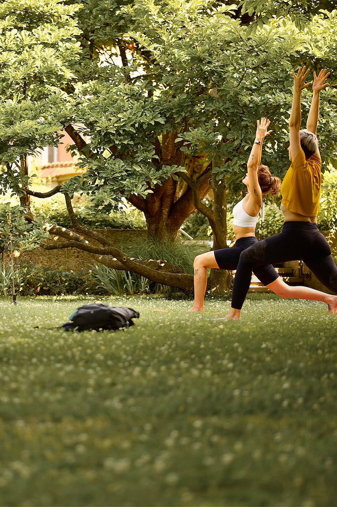 Zwei Yogaschülerinnen des LE MOUV Studios praktizieren die Krieger 1 Pose (Virabadhrasana 1) im grünen Hofgarten Augsburg, umgeben von idyllischer Natur.