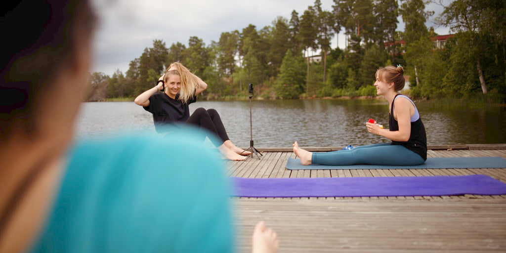 Leni Heim unterrichtet Yoga auf einem Holzsteg am See in Schweden. Eine Schülerin schaut lächelnd von der Seite zu ihr, während eine andere Schülerin frontal vor ihr sitzt. Natur, Ruhe und Vorfreude auf die Yogastunde sind spürbar.